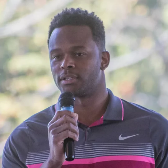 A man in a gray and pink Nike polo shirt speaking into a microphone with a blurred outdoor background.