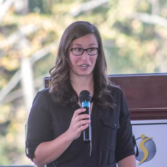 A woman with long brown hair and glasses speaking into a microphone, standing near a podium with a blurred outdoor background.