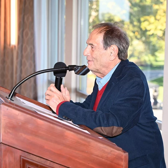An older man speaking at a podium with a microphone in a well-lit room with large windows overlooking greenery.