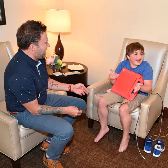 An adult man and a young boy sitting in chairs, laughing and pointing at each other, with the boy holding a red tablet in a cozy room with a lamp and decor.