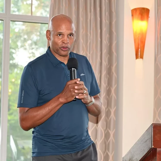 A man in a blue polo shirt speaking into a microphone in a room with large windows and warm lighting.