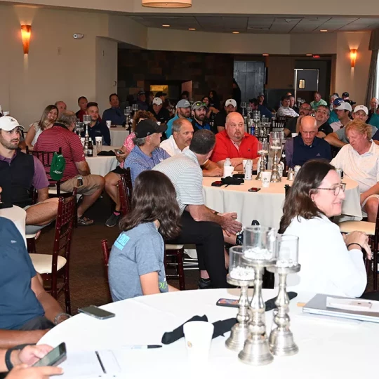 A large group of people seated at round tables in a well-lit banquet hall, attentively listening to something happening off-camera. The audience includes men and women of various ages, many of whom are wearing golf attire, suggesting they might be at a post-golf event gathering. The tables are set with decorative centerpieces, and the atmosphere appears social and engaged.