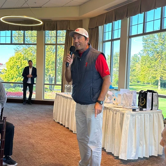 A man in a gray vest and red shirt speaking into a microphone in a bright room with large windows, with another person standing in the background near a display table.