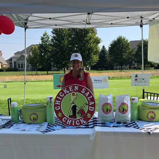 Smiling woman at a Chicken Salad Chick booth set up at an outdoor event.