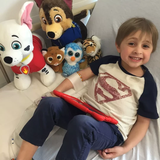 Smiling young boy in a hospital bed, surrounded by plush toys, holding a red tablet.