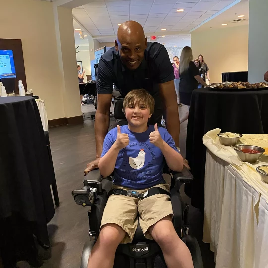 A young boy in a motorized wheelchair giving two thumbs up, with a smiling adult standing behind him in a room set up for an event with tables and food.