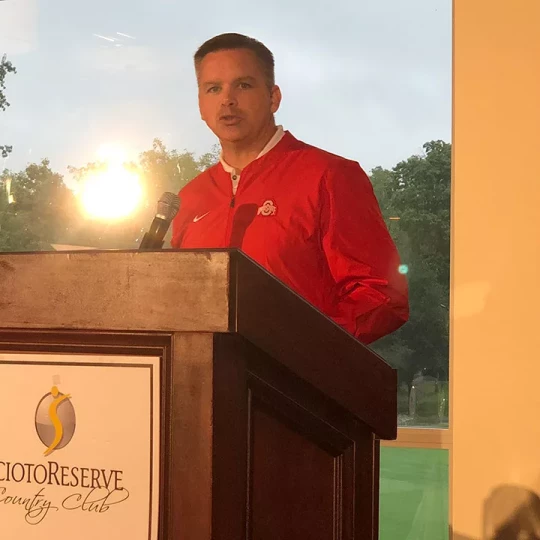 A man in a red Ohio State jacket speaking at a podium in front of a window with sunlight streaming in, with the Scioto Reserve Country Club logo visible on the podium.