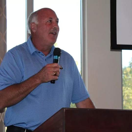 A man in a blue striped polo shirt speaking into a microphone at a podium in a room with large windows.