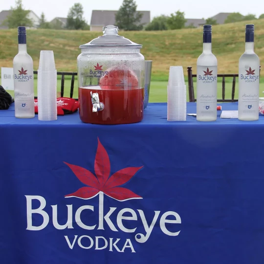 Table set up at an outdoor event featuring Buckeye Vodka bottles, a large dispenser of red punch, and plastic cups, with a blue tablecloth branded with the Buckeye Vodka logo.