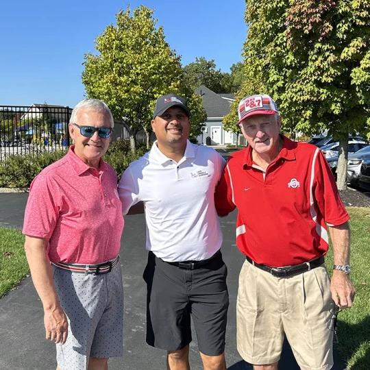 Three men standing together outdoors, smiling on a sunny day.