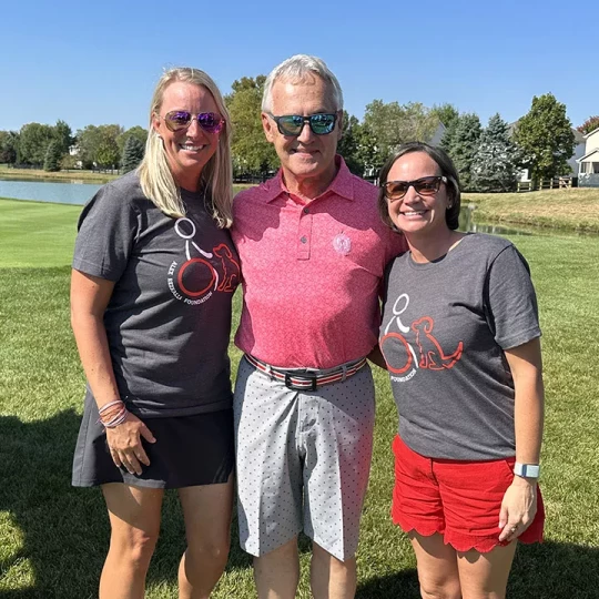 Three people posing together outdoors on a sunny day, two wearing foundation t-shirts and one in a pink polo shirt, with a grassy field and pond in the background.