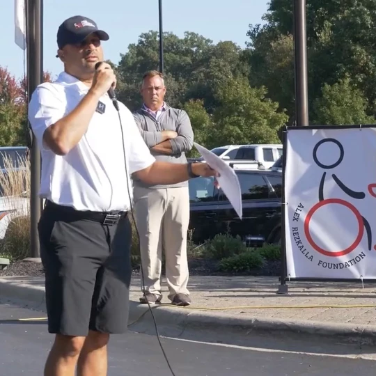 Man speaking into a microphone at an outdoor event, holding a paper, with a Alex Rezkalla foundation banner in the background.