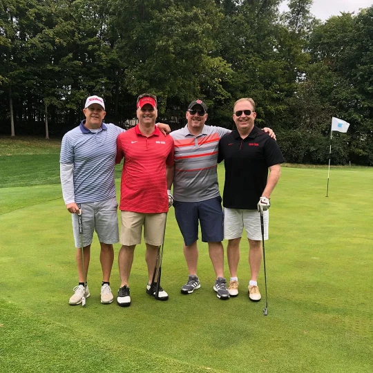 A group of four men standing on a golf green, smiling and posing for the camera. They are each holding golf clubs, with one man wearing a red Ohio State shirt and the others in a mix of golf attire. The setting is a golf course with trees in the background.