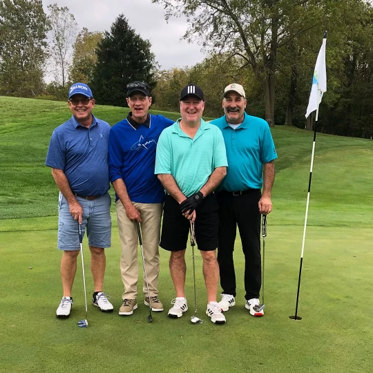 Four golfers standing together on a green course, each holding a golf club, with a flagpole and hole nearby. They are dressed in casual golf attire with smiles, enjoying a day on the course.