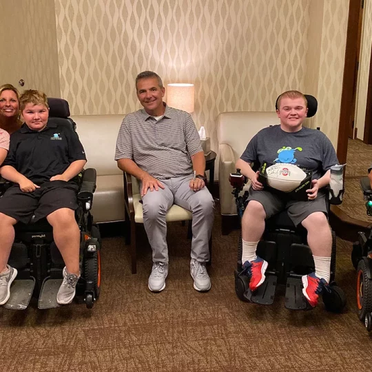 A group photo in a room with a man seated in the center, surrounded by two young boys in motorized wheelchairs, one holding a signed football.