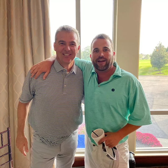 Two men standing indoors, smiling with arms around each other.