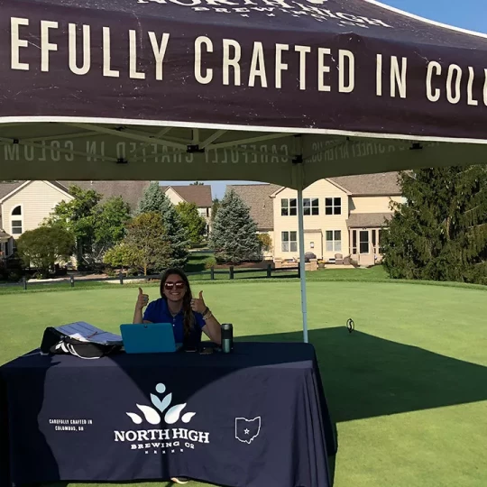 A woman sitting at a table under a North High Brewing tent at an outdoor event, giving two thumbs up, with houses and trees in the background.