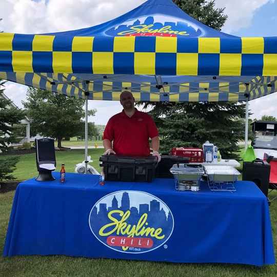 A man standing behind a table under a Skyline Chili tent at an outdoor event, with food and supplies set up on the table.