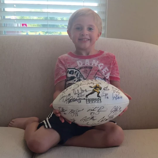 Smiling young boy sitting on a couch, holding a football signed by Heisman Trophy winners.
