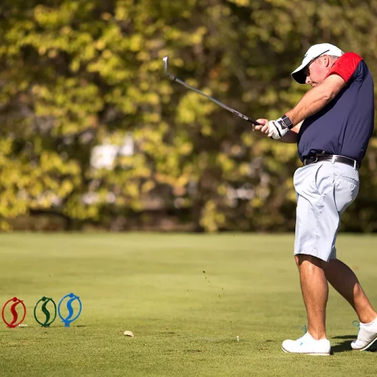 A golfer in mid-swing on a green course, wearing a navy and red shirt, light blue shorts, and a white cap, with trees blurred in the background.