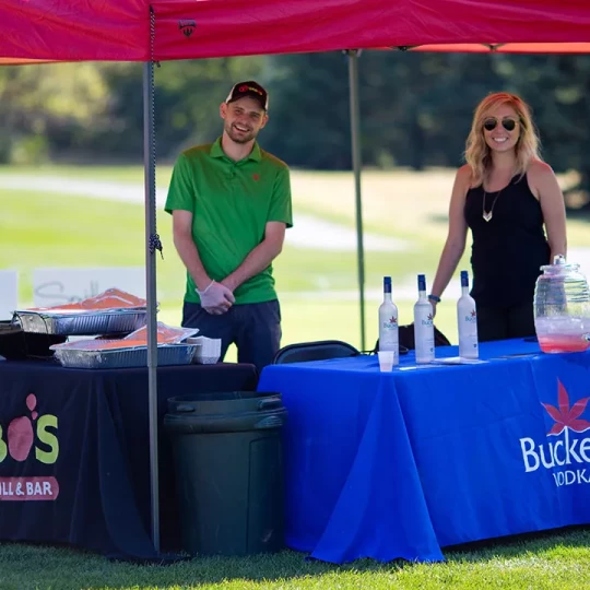 Two people standing under a tent at an outdoor event, with food and beverage tables set up.