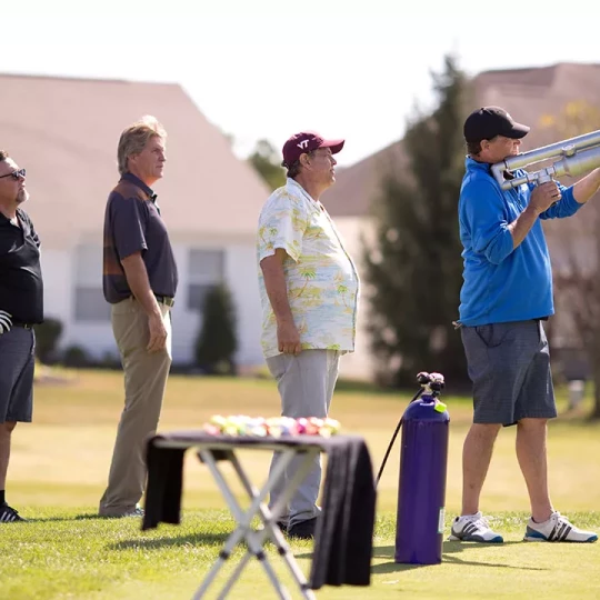 A group of men standing on a golf course, with one man in a blue jacket holding a large air cannon, preparing to launch a golf ball.